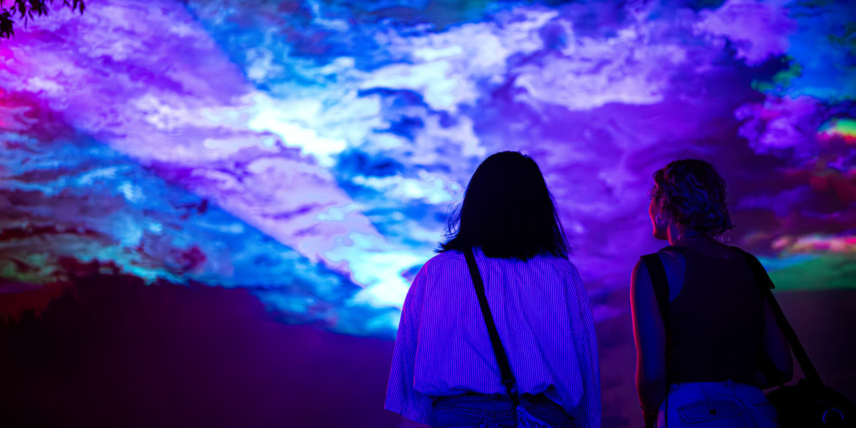 Two young women gaze upwards toward a clouded sky filled with the colours of Borealis.