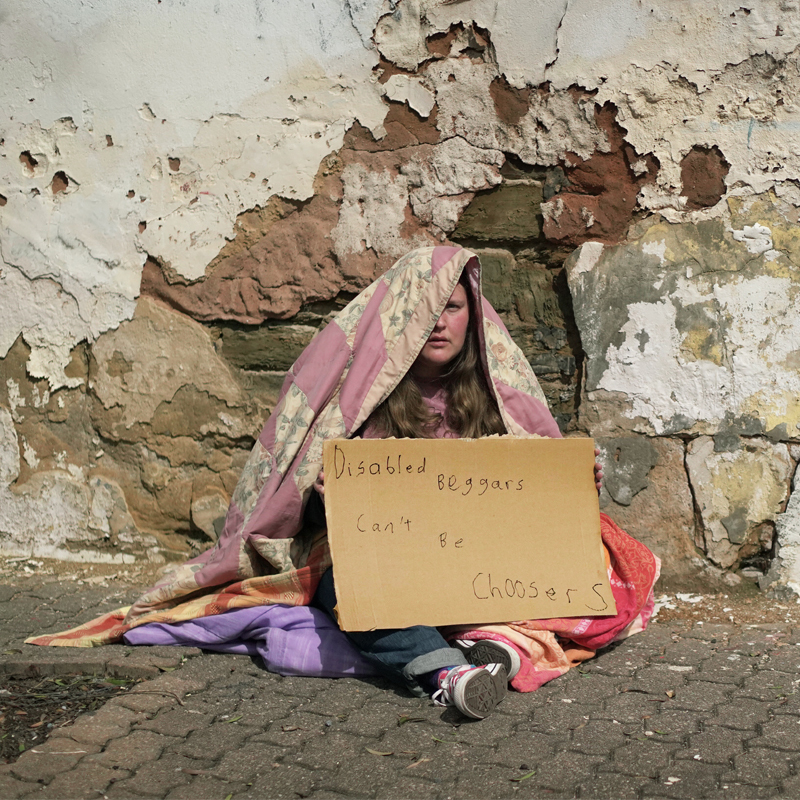A woman is sitting on the ground wrapped in blankets. She is sitting in front of a stone brick wall. She is wearing a pink long-sleeved t-shirt jeans and pink trainers. She is holding up a cardboard sign saying "Disabled Beggars Can't Be Choosers"