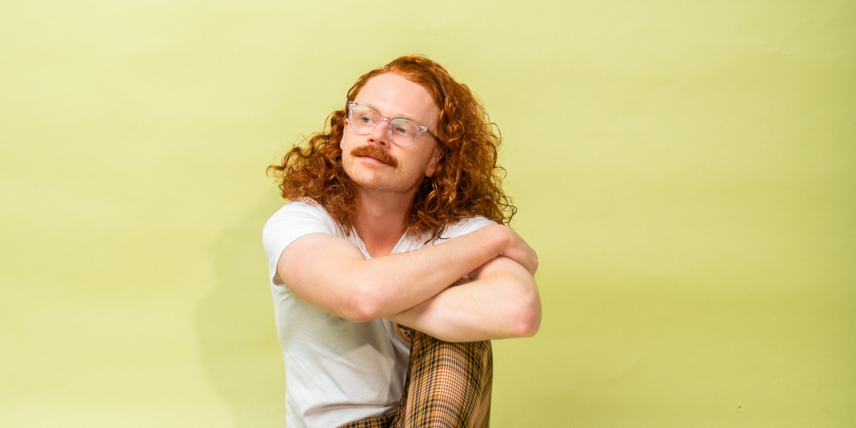 Nick sits with his arms crossed over one knee in front of a light green backdrop with his long curly red hair out. He's wearing orange and brown checked pants and a white tee shirt. He's starting inquisitively off to the right.