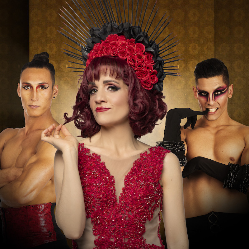 Rouge - Woman stands central in a red dress with a red and black spiky headdress. She twists a lock of hair in her fingers and looks at the camera with a coy look on her face. Two muscled shirtless men flank her, the one on the left has a serious look on his face with his arms folded. The man on the right has theatrical drag style make up, a cheeky grin, and long black silk gloves.