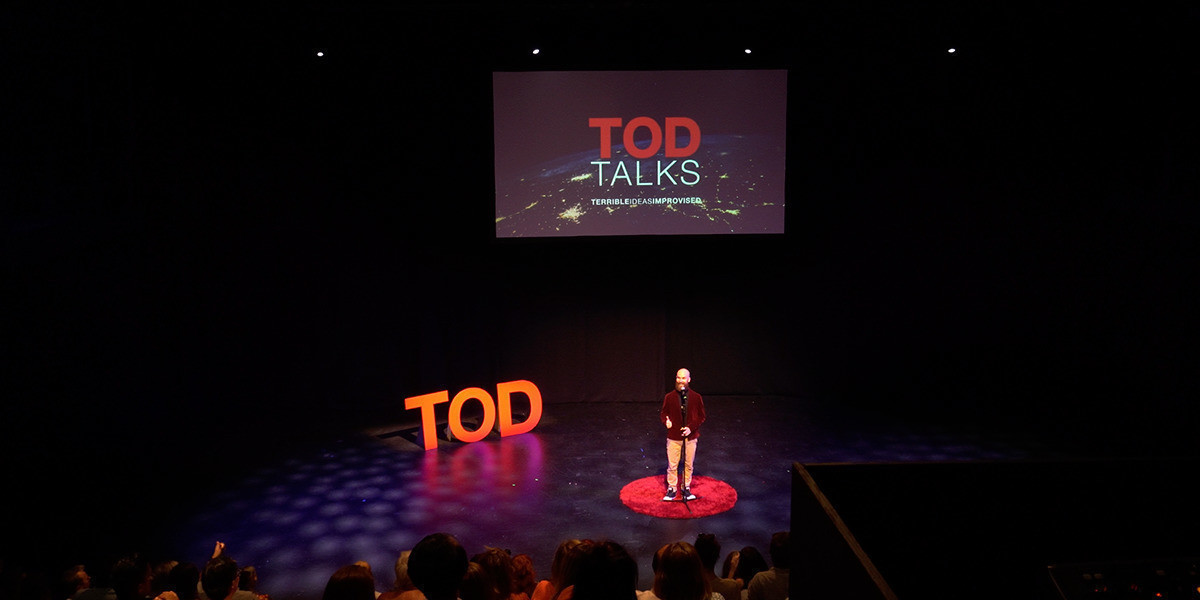 A presenter on a stage with an overhead projector, a red round carpet, and 3 cutout letters that say TOD.