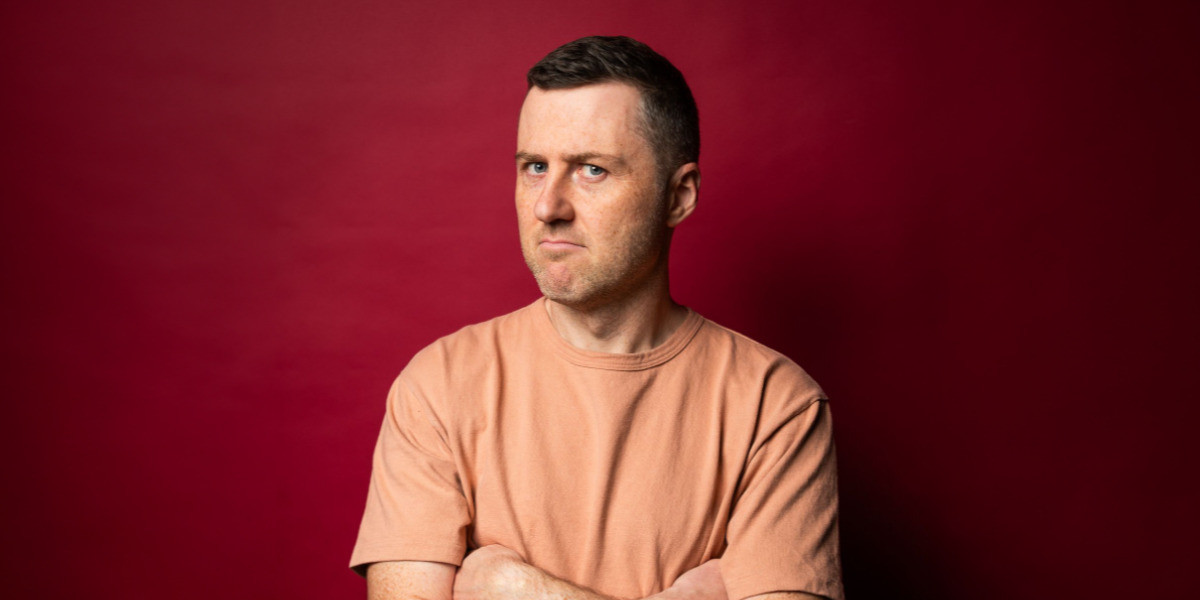 Lloyd’s head is tilted to the side but his eyes are looking directly into the camera. He has his armed crossed and he has an annoyed expression on his face. He is wearing an orange tshirt against a red backdrop. He has short brown hair and blue eyes.