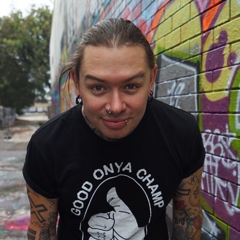 A photo of a man with multiple tattoos on his arms wearing a black t-shirt with an examining facial expression. He has multiple black earrings in his ears as well as a silver piercing on his nose and lip. The background features a brick wall covered in colourful graffiti.