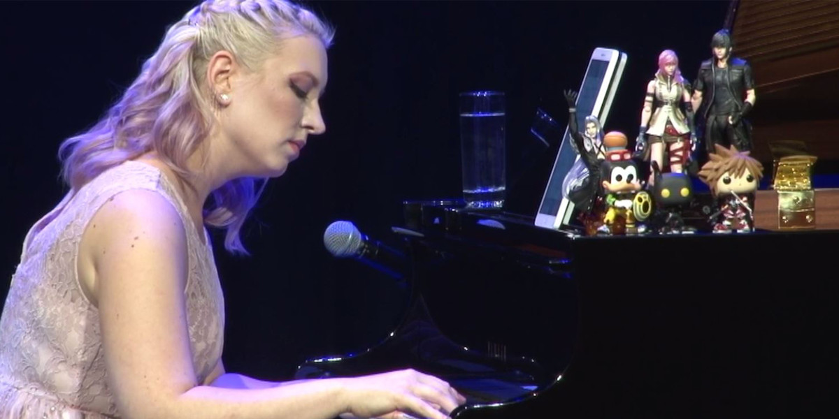 blonde woman with wavy hair is playing the piano with a serious expression on her face. she is wearing a pink feather dress and there are video game figurines on top of the piano