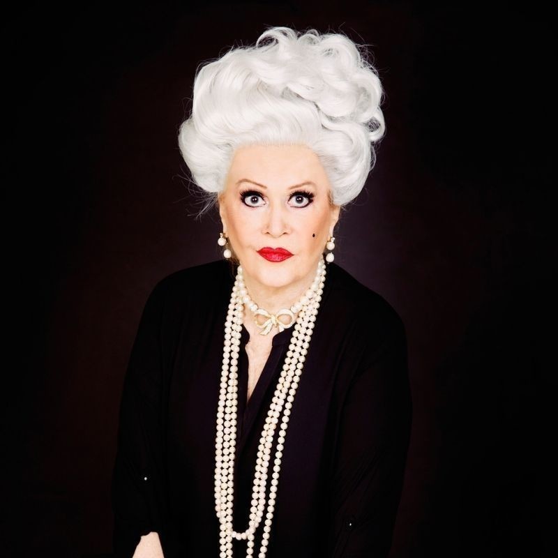 Carlotta: I'm Not Dead Yet Darlings! - A photograph of a person wearing a large grey wig and red lipstick. They are wearing a long sleeved black shirt and multiple pearl necklaces around their neck.