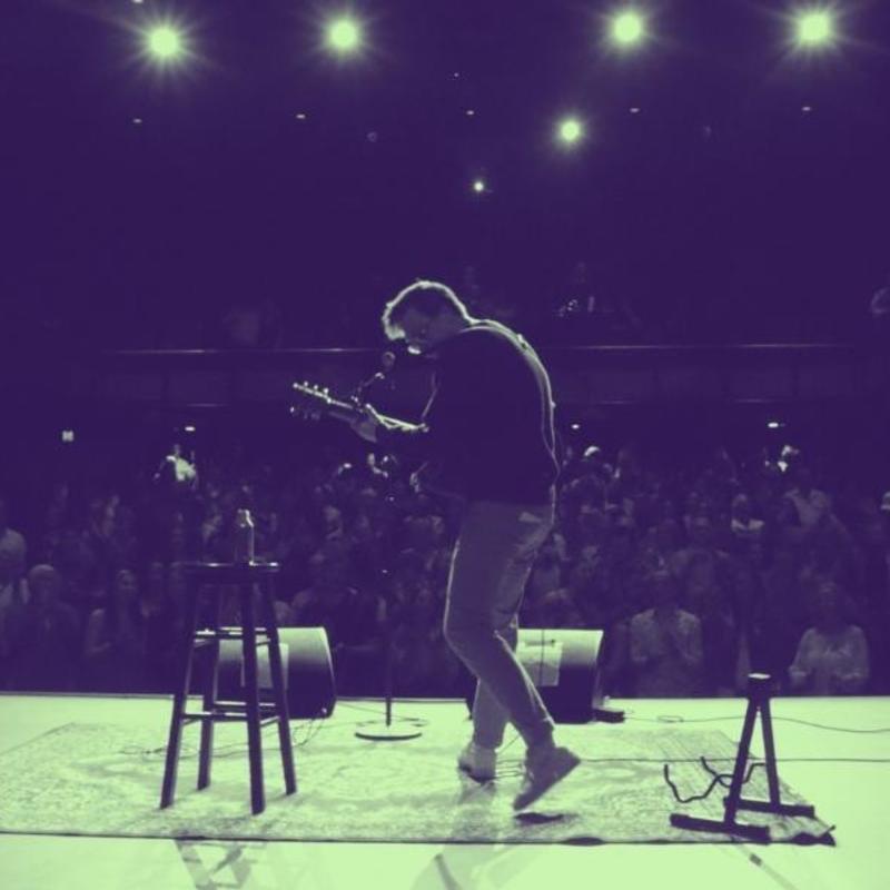 Comedian Peter James, on stage in a theatre playing an acoustic guitar, taken from the perspective of behind the performer, facing the audience.