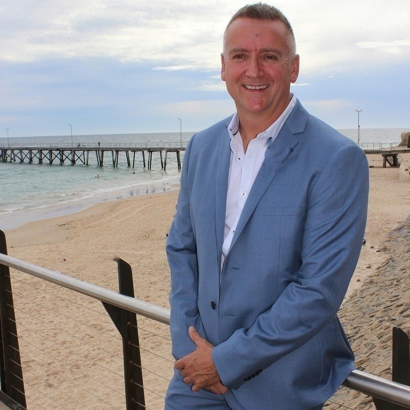 A man in a light blue suite is leaning against a fence. Behind him is the beach and a jetty. It is an overcast day.