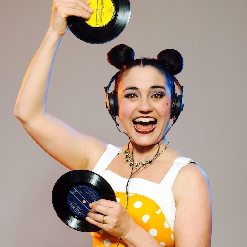 Broadly smiling performer, Monski Mouse holds two 45' vinyl records, one above her head and one in front of her, she has her hair fashioned into two big buns on her head, made to look like mouse ears, and where's a yellow and white polka dot 1950's style swing dress.