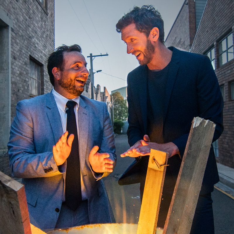 A RATIONAL FEAR - LIVE - Image from left to right: Dan Ilic has short brown hair and a beard. He is wearing a grey suit, white collared shirt and dark tie. Lewis Hobba has dark short hair and a beard. He is wearing a dark shirt and a navy blue dinner jacket. Both men are standing over a bonfire and looking at each other smiling. They are standing in an industrial looking alleyway.