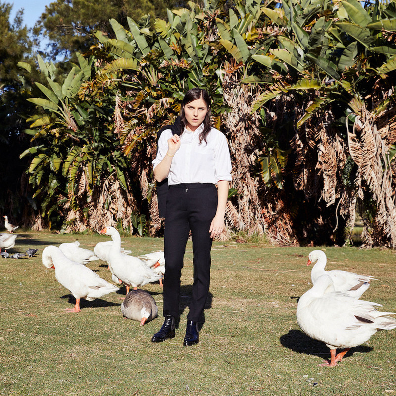 Ange Lavoipierre: I’ve Got 99 Problems and Here Is An Exhaustive List Of Them - Ange stands on grass, in front of tress wearing a white shirt. There are swans milling around her feet in both the left and right corners of the shot.