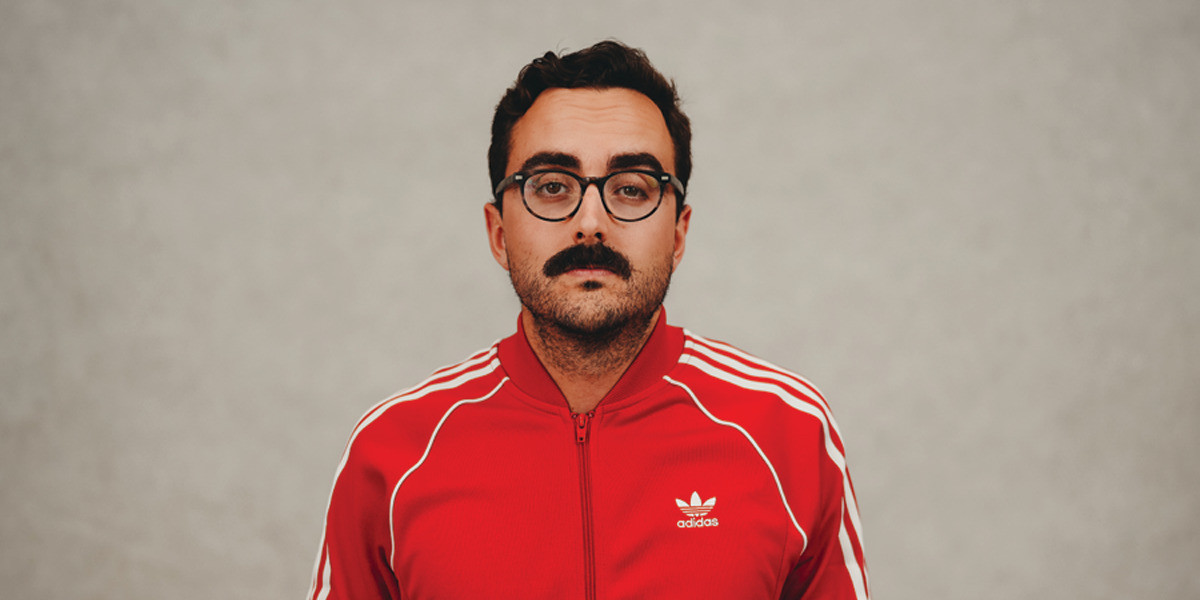 White man, dressed in red and white adidas tracksuit, stares at the camera with a deadpan expression. He has glasses and a moustache.
