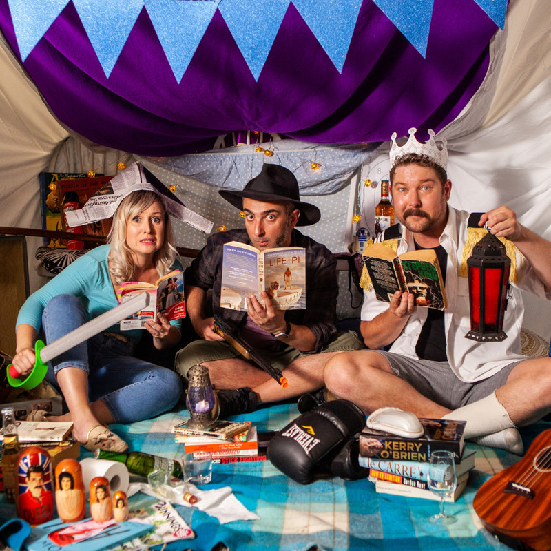 Three actors sit in a homemade blanket fort. They are holding books and swords and two are wearing paper hats and crowns.