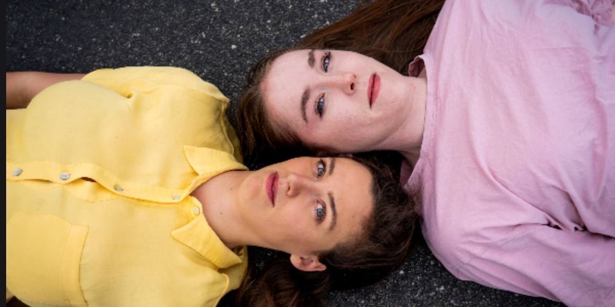 Two young women lie on the ground looking up at the sky, their heads close together. One is wearing yellow, the other pink. Their expression is one of defeat.