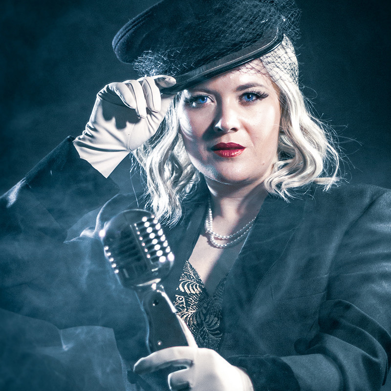 A woman with white hair in a cabaret costume holds a vintage microphone with a white gloved hand. The woman is tipping her military-style hat and is surrounded by theatrical smoke.