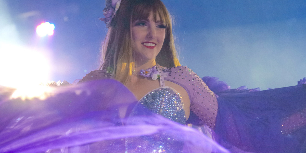 A close up image of a Female, Blonde Burlesque Performer, dressed in a glamorous purple costume, dancing on stage. The stage lighting and haze can be seen in the background of the image.