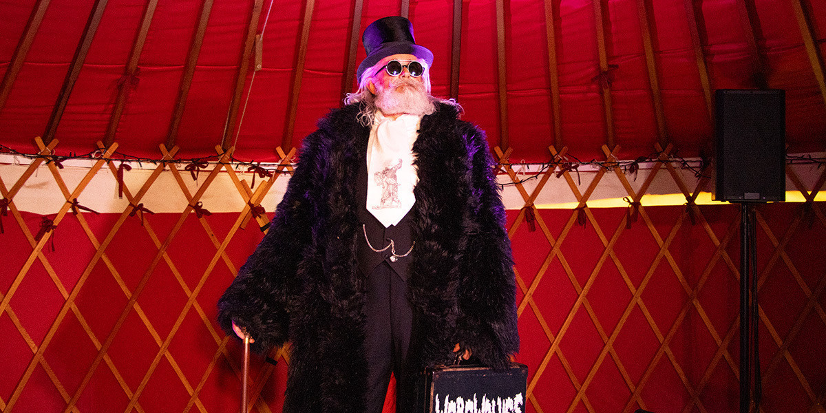 A gentleman stands inside a red lined yurt with the dark wooden roof beams of the structure showing against the red cloth. He has long grey hair and neatly trimmed beard with a curled moustache. He is wearing round dark glasses. He is dressed all in black save for a white shirt and cravat with an old 18th century print of a werewolf devouring a villager. He is wearing a huge black fur coat with a top hat, walking cane and a case with 'Werewolves' written on it.