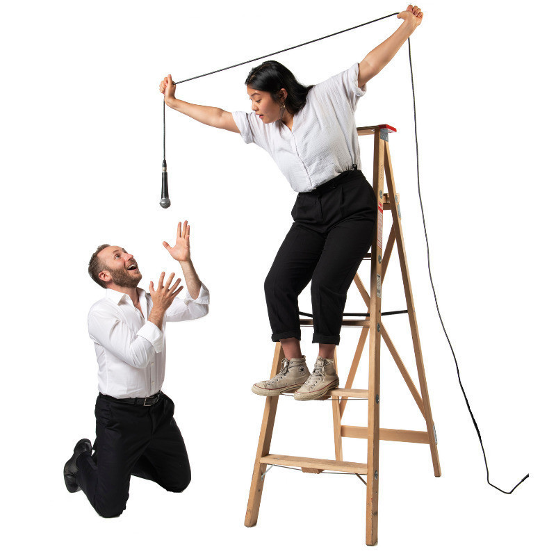 Woman standing on a ladder holding the microphone above a mans head as he sings to it while kneeling down. Both are dress in a white top and black pants.