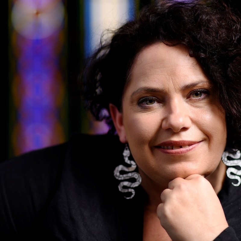 Performer Nancy Bates is seated in front of a church's stained glass window with beautiful colour shining in the background.  A warm small smile on her face, wearing a black shirt, and long black and white snake earring with an Indigneous design.