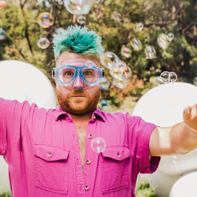 A performer is wearing a bright pink shirt and snorkling goggles. He is surrounded by bubbles