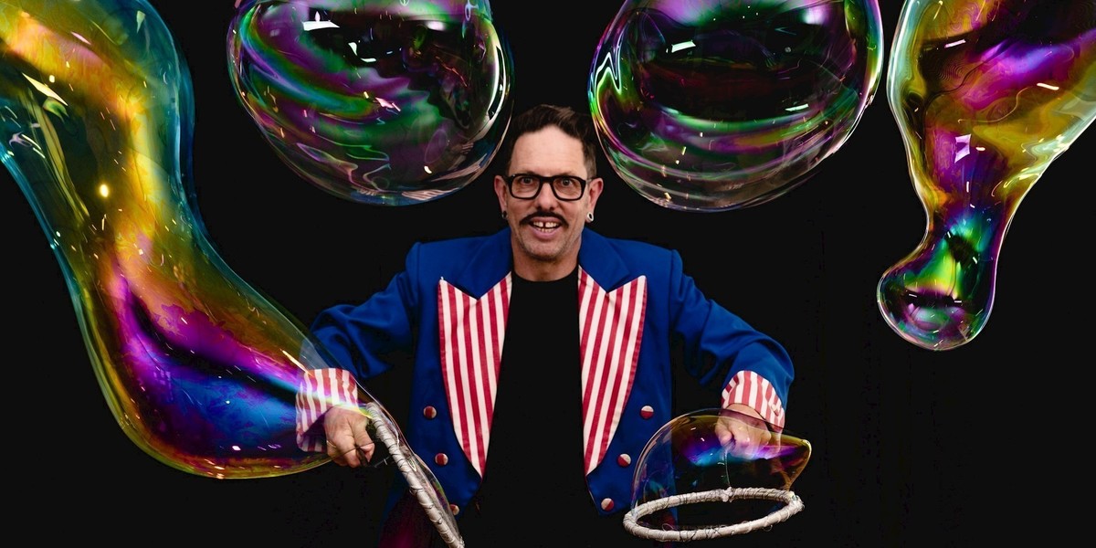 A man with glasses and a moustache in a blue jacket with red striped lapels, stands in front of four giant oblong shaped bubbles.