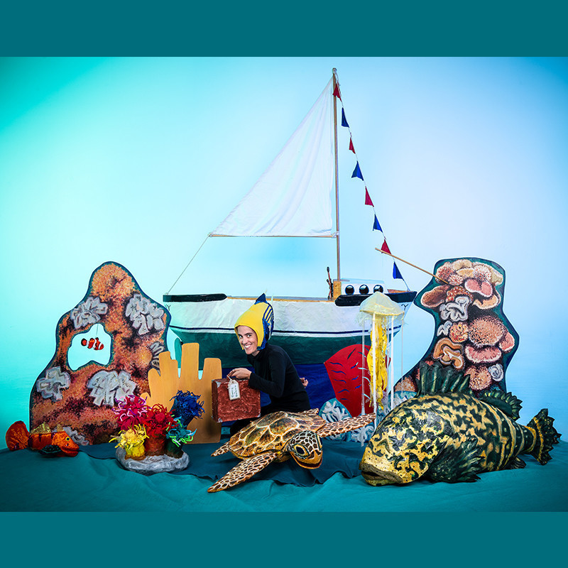 The puppeteer is dressed like a fish in front of a sailing boat. Surrounded by the coral reef, a sea turtle, grouper fish, and jellyfish.