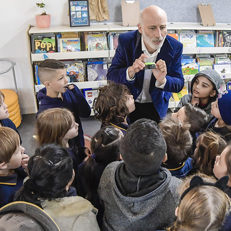 Stephen, man in 50s, holding blue button, surrounded by primary school students in a school library.