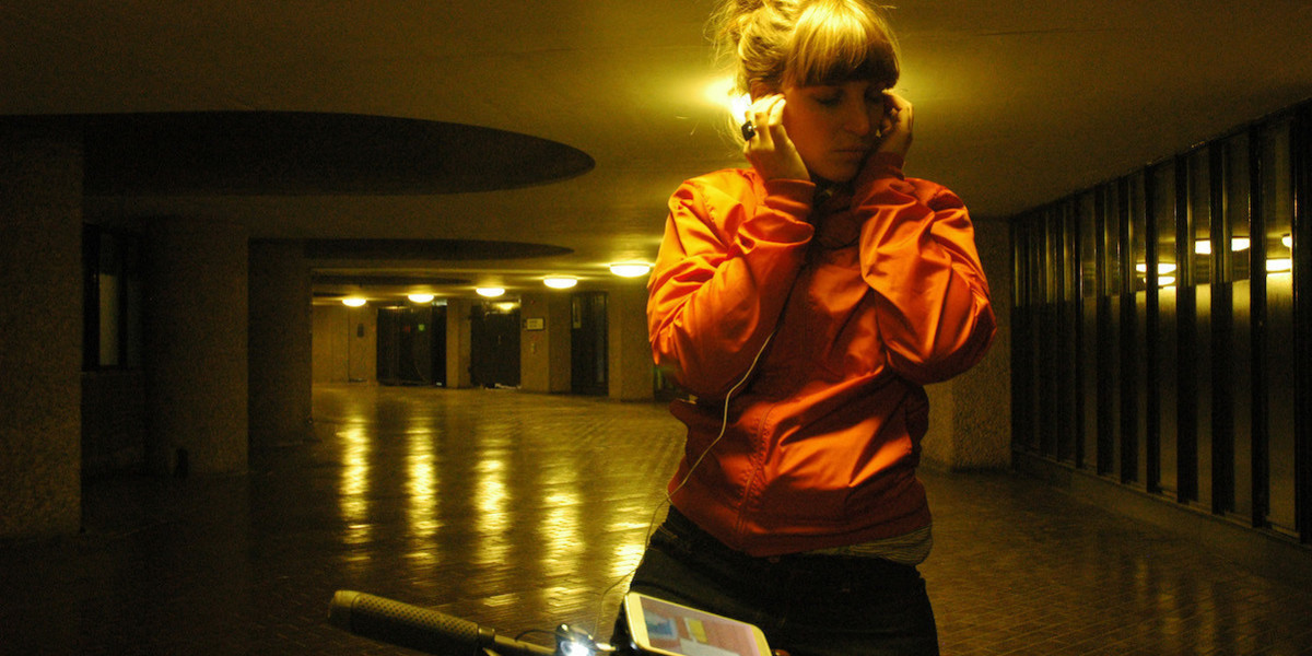 A person standing next to a bike, wearing an orange jacket, looks down at a smartphone mounted on their bike, with their left hand holding headphones to one ear.