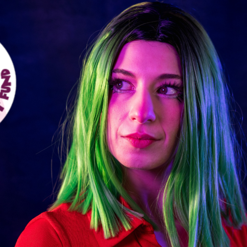 A performer in a red top and green, shoulder-length wig looks off-camera. She has a wistful expression on her face. There is a circular Supported by the Adelaide Fringe Artist Fund logo.