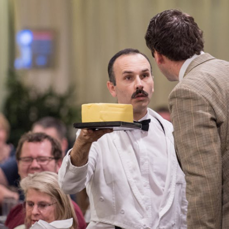 A waiter is carrying a tray of butter and appears with a confused expression on his face. Another man is facing towards him.