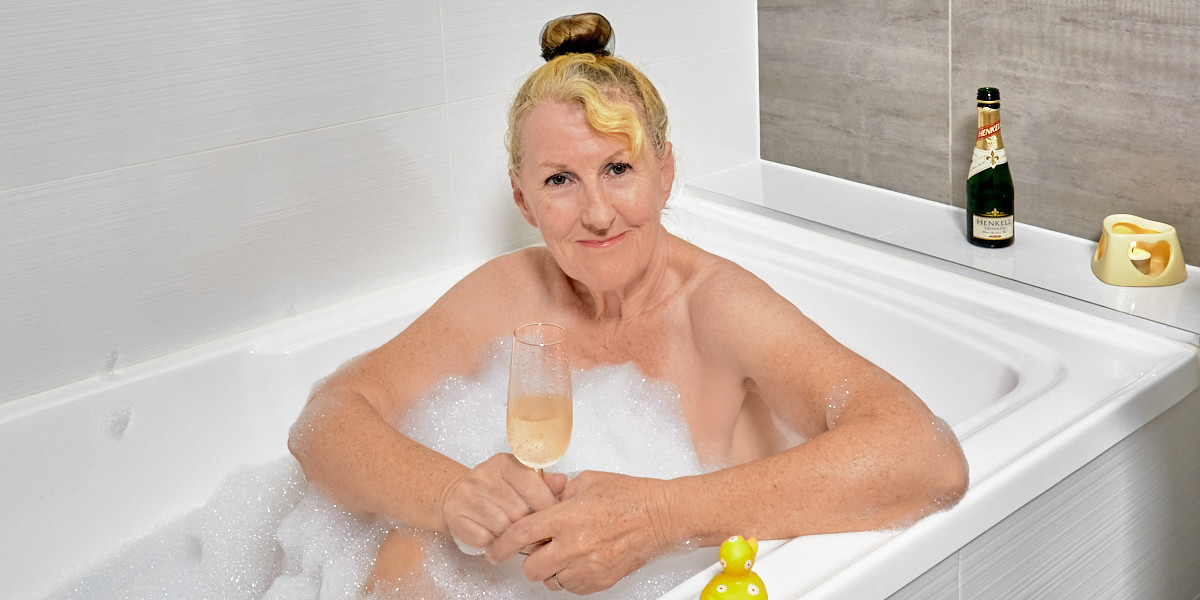 The presenter is in a bubble bath, holding a glass of sparkling wine