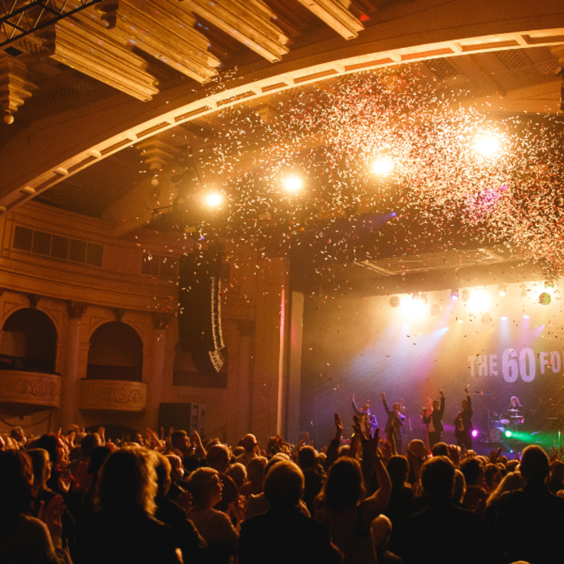 The Thebarton Theatre washed with striking gold lights, as confetti rains down inside the auditorium. In the foreground, the audience are raised in a standing ovation, with four performers on the stage in a final pose with their hands in the air.