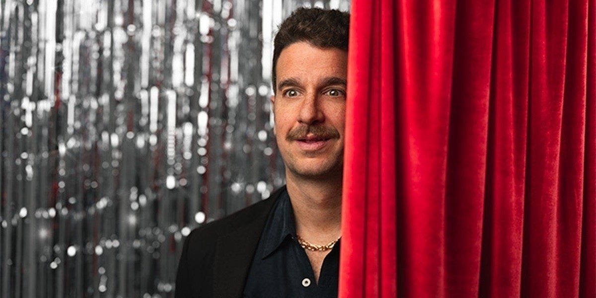 Mike stands behind a red velvet curtain in the foreground with a silver party curtain background.