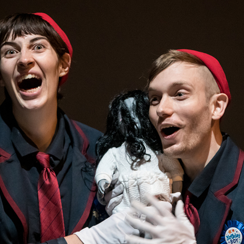 The two creepy boys are holding a child sized doll with long black hair. Both boys are dressed in dark blue blazers (similar to a child's school uniform).  They both are wearing small red touques, and white gloves.  They both have red ties.

The doll has hair covering her face, is dressed in a white dress, and has grey skin.

The boy on the left (Grumms) has dark hair, and the boy on the right (Sam) has light brown hair.