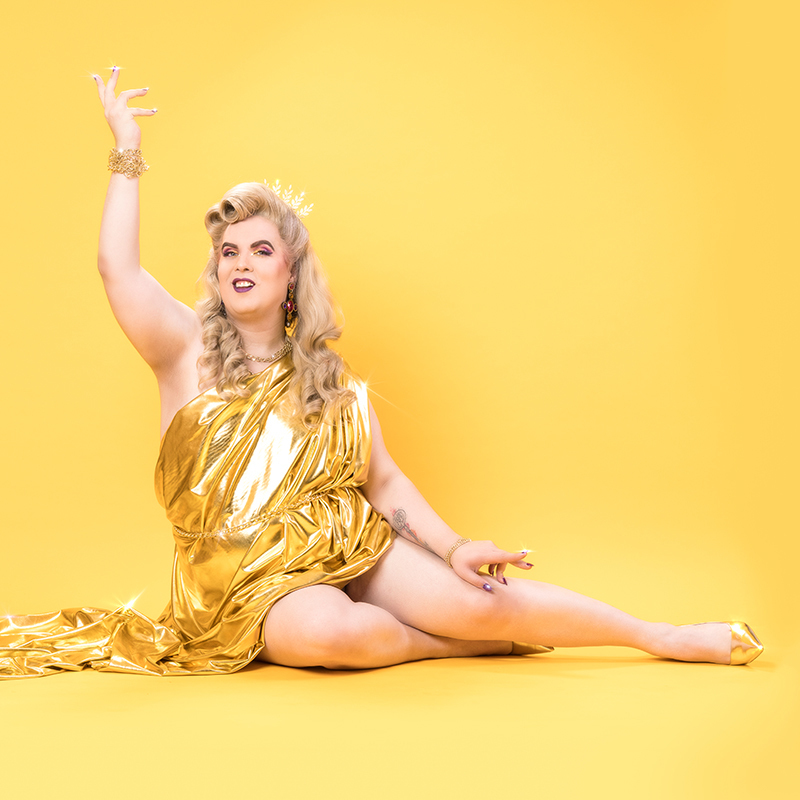 Anna Piper Scott: Such An Inspiration - Anna Piper Scott, a stunning trans woman, reclines on a bright yellow backdrop. She has blonde hair, purple nails and is wearing gold shoes, a gold tiara and gold dress, which drapes across the floor next to her. She has a confident smile, and an arm raised in a flamboyant pose.