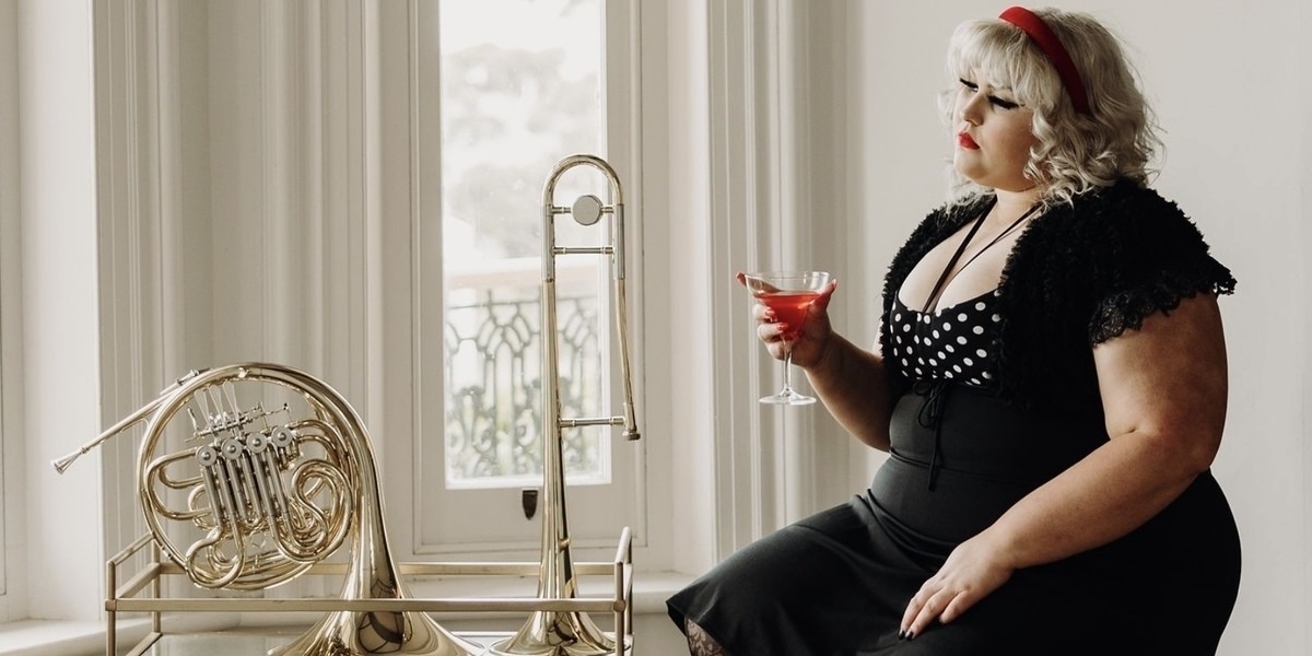 Charlee Watt, a young woman with platimum blond hair, big black eyeliner and lashes, and bold red lips, wearing a black dress with white polka dot bodice, holding a red cocktail drink looking out of the window. Beside her is a vintage glass and gold drinks cart upon which is placed an array of brass instruments.