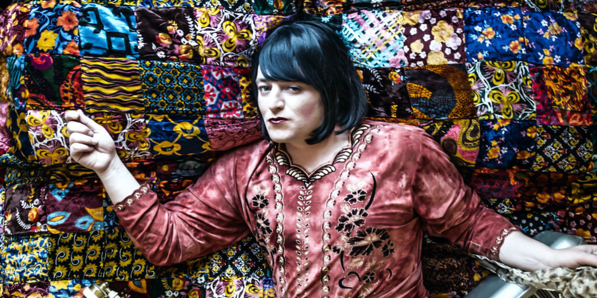 Singer with a black bob lying on a bed with a colourful bedspread and items such as a piano accordion, old wired telephone and pillows.