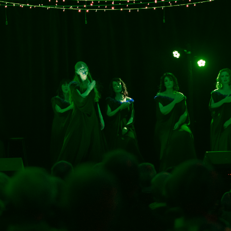 A group of singers sits in a semi-circle looking towards a soloist standing in the centre facing the audience. All singers are holding their right hand over their heart.