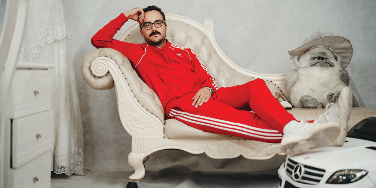 Daniel Muggleton, dressed in red and white adidas tracksuit, reclines on a white chaise lounge with his head in feet on a tiny white Mercedes.