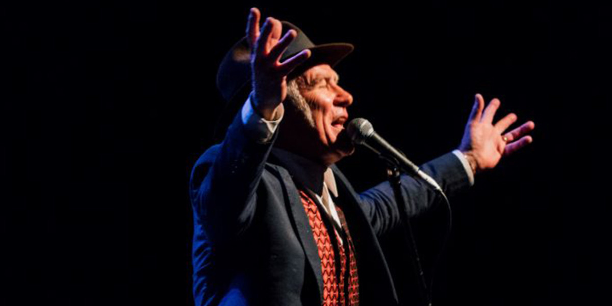 Stewart D'Arrietta is wearing a blue suit, crisp white shirt and an embroidered red satin vest, as well as his signature hat, standing at the mic, facing right. Both his arms and all his fingers are outstretched above his head, and his faces is lifted, eyes closed, reaching out to the heavens as he belts one out.