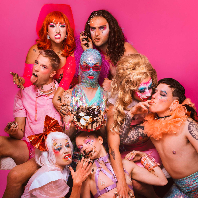 A picture of 8 artists on a pink background. The top left artist has long red hair and is snarling, the top right artist has long brown hair and is looking directly into the camera. The middle left artist has short hair, wearing a pink shirt, and is poking their tongue out. The middle artist is wearing a face-kini that is incrusted with gems, with a pair if red sunglasses over the top. The two artists on the right hand side are leaning in towards each other intimately. One has full pink, white and blue face paint on, and the other is sucking on one of their fingers. The two artists on the bottom are leaning against one another, one with a pink wig and red bow, and the other dressed in a pink harness.