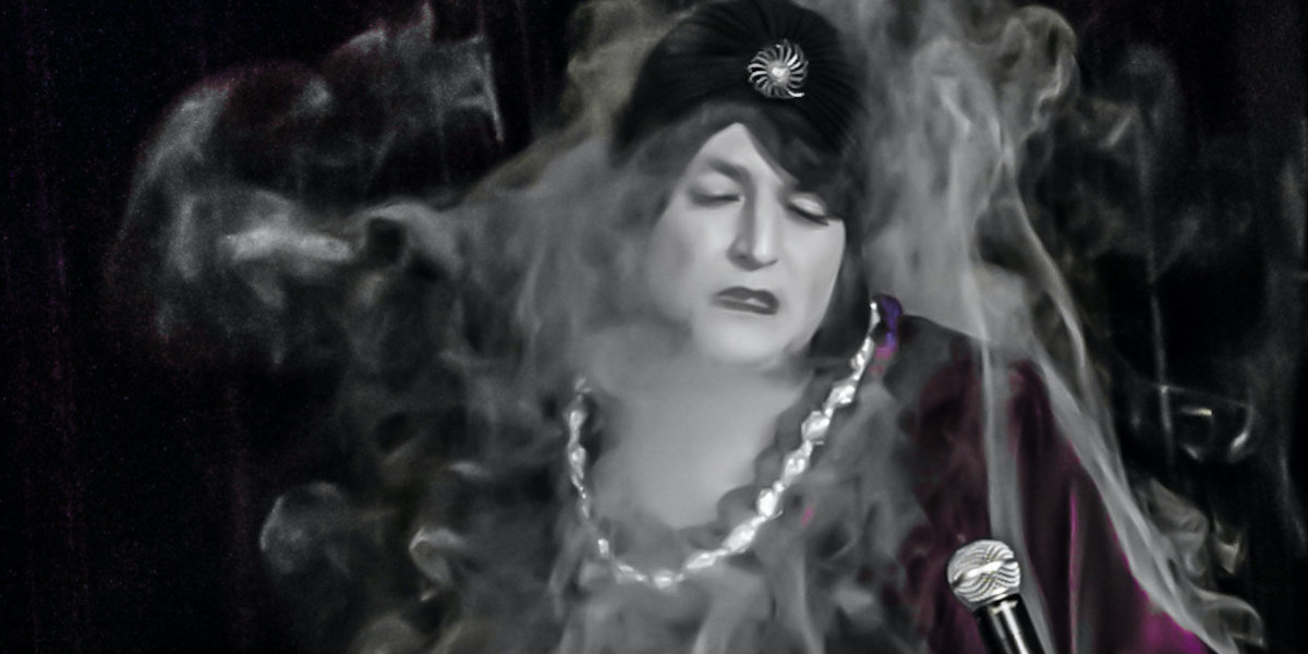 Singer in smoke, eyes closed, face turned toward microphone in their hand, surrounded by smoke. They have a white shell necklace on and 1920s style outfit on.