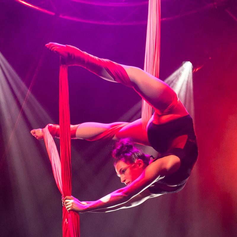 Performer from YUCK Circus is suspended in the air in a backbend position, hanging from a red silk. She pulls her feet towards her head, lit by rock and roll red lighting