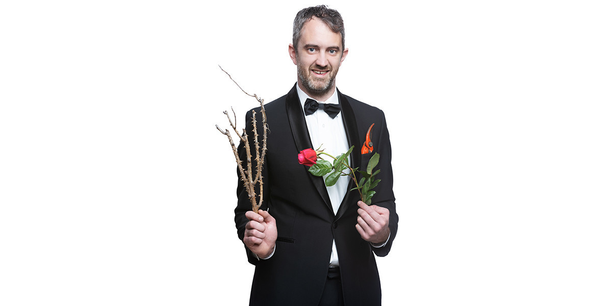 Kel's wearing a tuxedo holding a wilted rose in one hand and a thorny dead rose bush in the other. Pair of secateurs in his pocket.