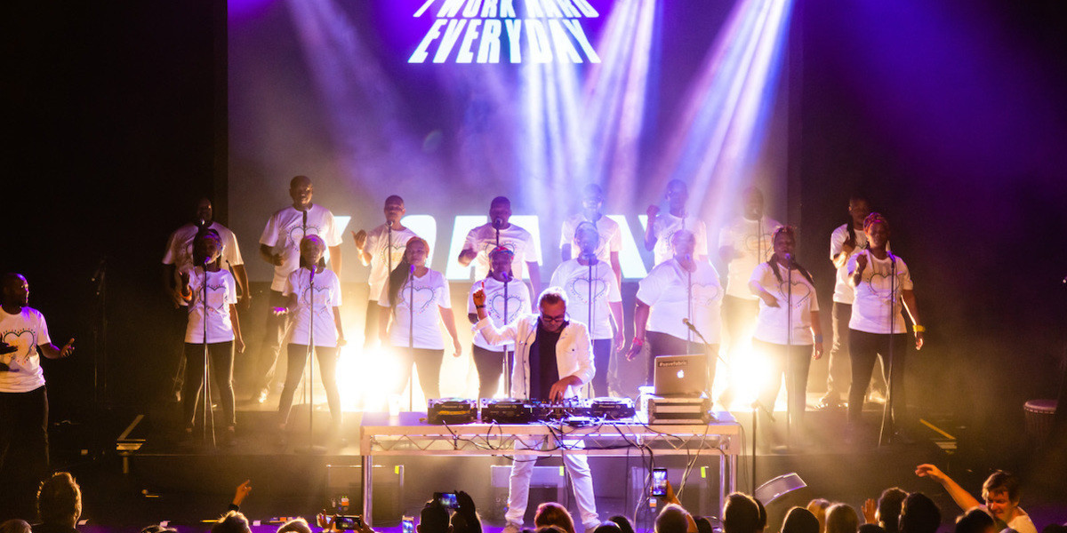A DJ, Groove Terminator, stands in front of Soweto Gospel Choir, with one fist in the air and the other spinning a record. Bright white, yellow and purple lights shine down on them. A silhouette of the audience is at the bottom of the image.