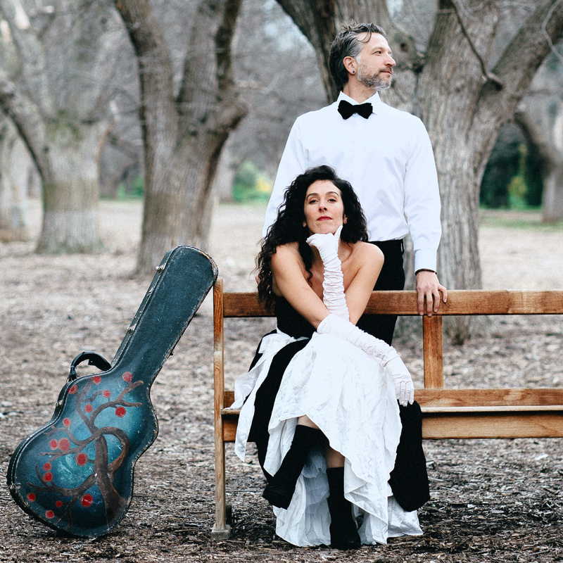 Ephemera - A woman with long dark hair wearing a black and white dress sitting on a bench amongst brown leafless trees. A man is standing close behind her looking away, he's wearing a white shirt and bow tie. A guitar is leaning against the bench beside them.