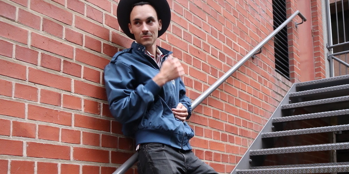 Comedian Andrew Silverwood in a Blue flying jacket and grey hat leans against a wall with a slight smirk on his face