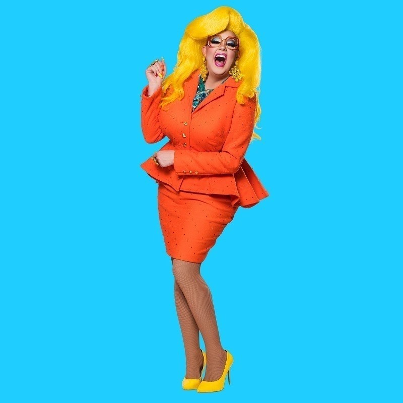 Karen From Finance - Karen From Finance is looking at the camera laughing. She is wearing a bright orange skirt suit with yellow hair and shoes in front of a blue background.