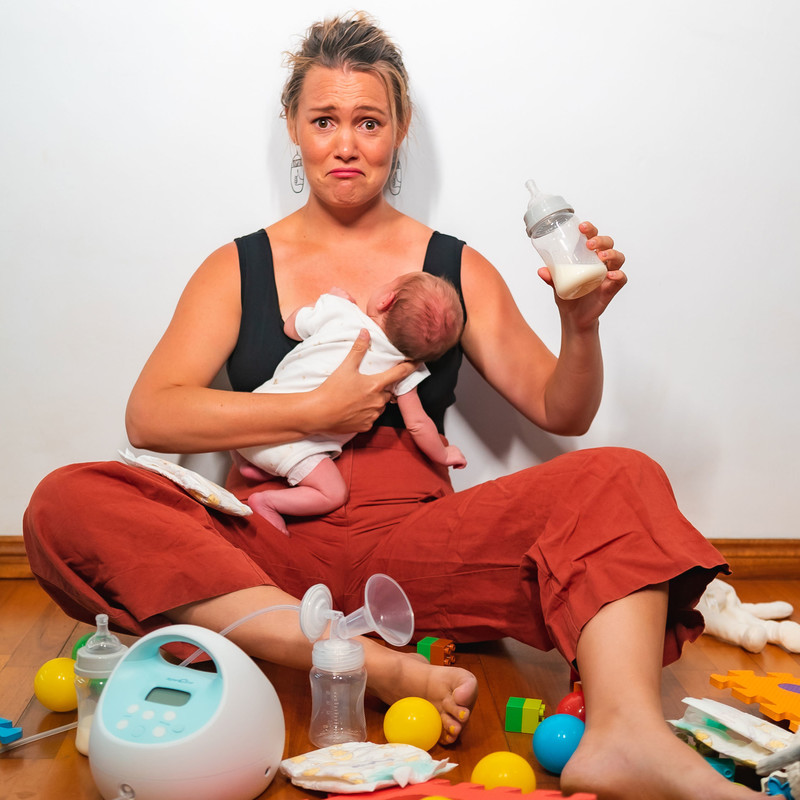A girl sits on the floor holding a baby and a bottle. Surrounded by toys and breast pumps and newborn baby items. She looks surprised.