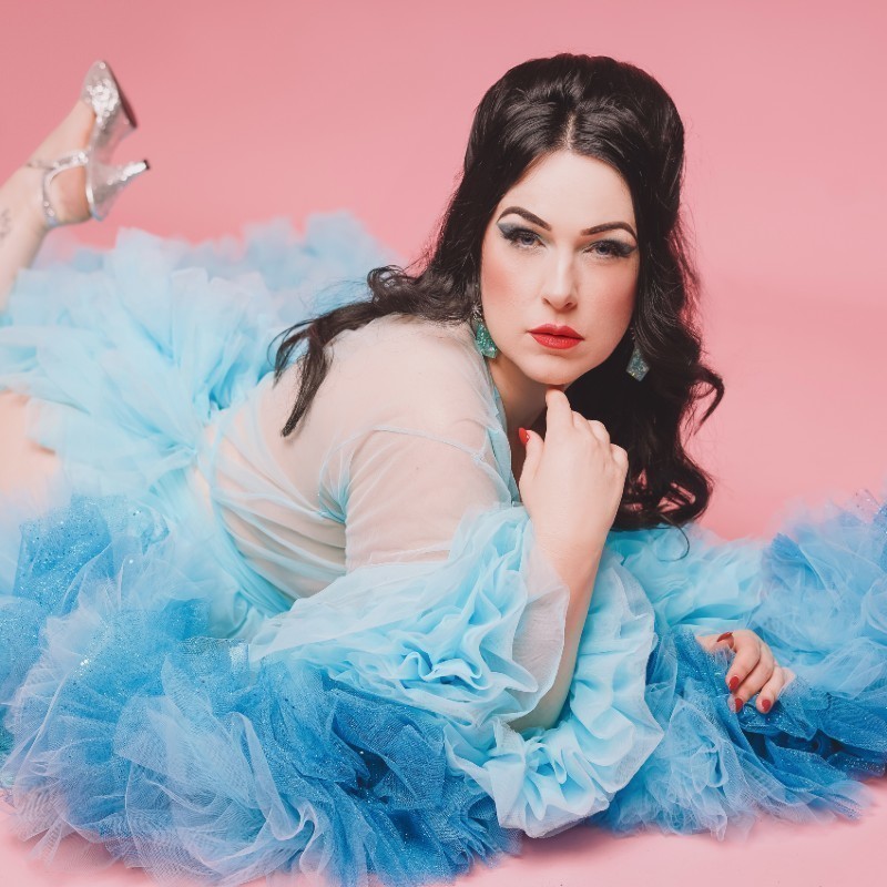 Brunette Burlesque performer Delza Skye lies on the ground, dressed in a light blue robe, with 1960s styled hair and makeup.