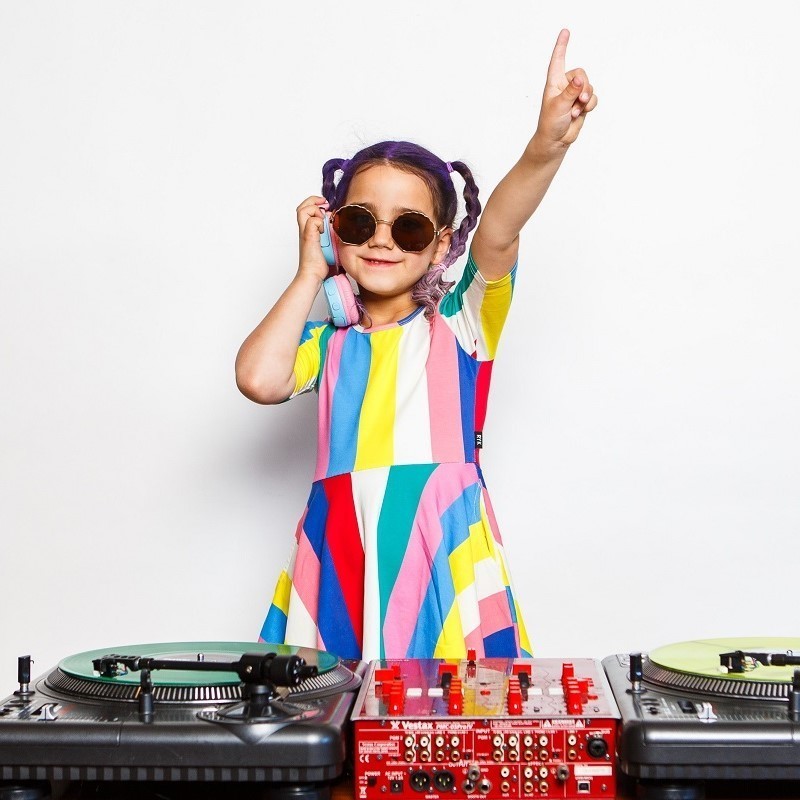 A kid in a colourful dress with purple pigtails is spinning some vinyl records on some decks